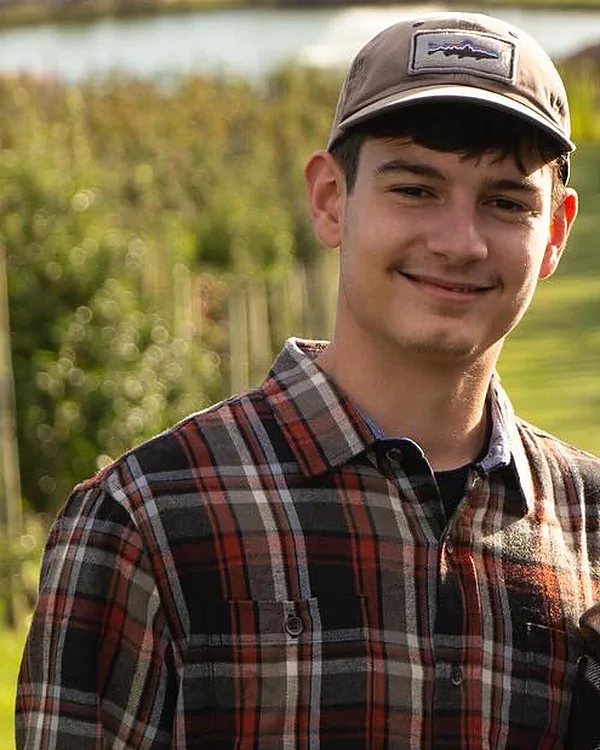 Nate Hammes, owner of Hammes Tree Service, smiling in a cap and flannel shirt with orchard rows behind him